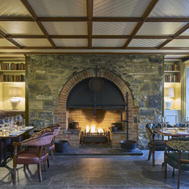 Cosy dining room with a lit stone fireplace, set tables, and warm ambient lighting.