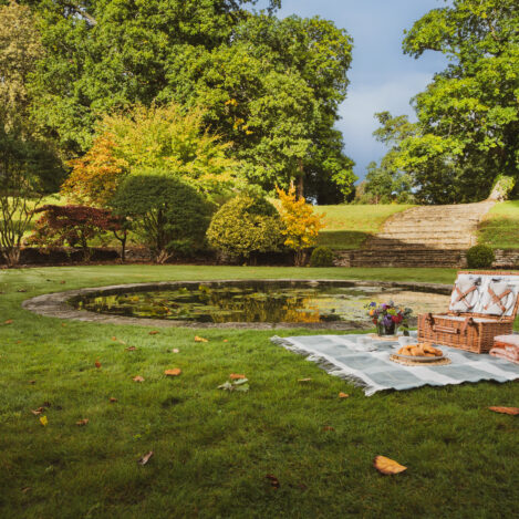 Cosy picnic by a pond in a lush garden, with a basket and blanket under sunny skies.