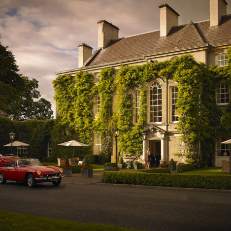 Elegant manor hotel entrance with ivy-covered facade; couple arriving in red convertible.