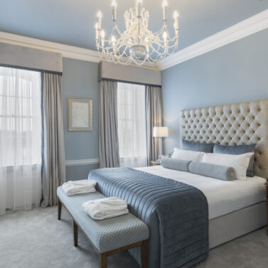 Elegant hotel room with king-sized bed, plush blue accents, and a chandelier.