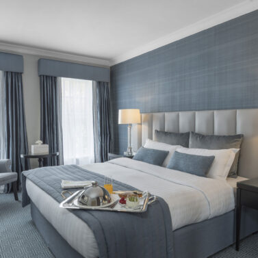 Elegant hotel room with breakfast tray on bed, soft lighting, and cosy seating.