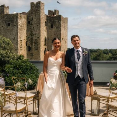 A joyful couple in wedding attire walking hand-in-hand near a historic castle on a sunny day.
