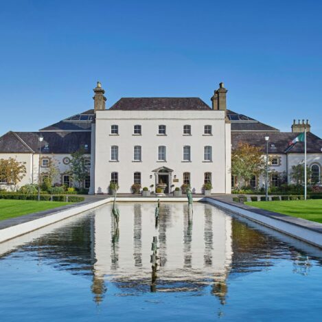 Elegant hotel building with a reflecting pool, surrounded by lush green lawns under a clear blue sky.