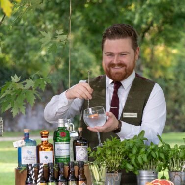 Friendly bartender mixes drinks at outdoor bar surrounded by lush greenery.