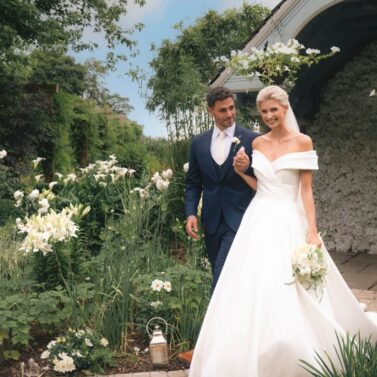 A joyful couple in wedding attire walks through a lush garden, surrounded by blooming flowers.