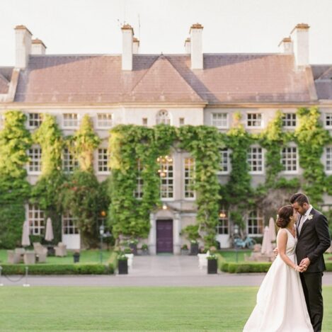 A couple embraces on a lawn in front of an ivy-covered, elegant hotel.