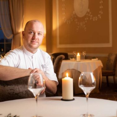 A chef warmly welcomes guests at a cosy, candlelit dining table in an elegant restaurant.