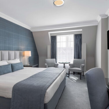 Inviting hotel room with a king bed, soft grey and blue decor, and a sitting area by a window.