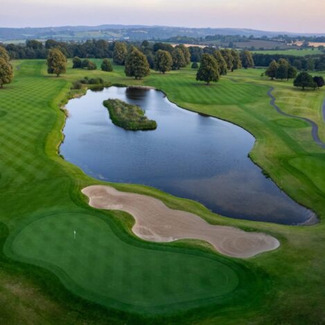 Aerial view of lush golf course with serene pond, well-maintained greens, and surrounding trees.
