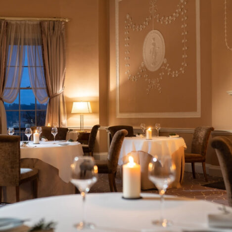 Elegant dining room with candlelit tables, a roaring fireplace, and large windows at dusk.