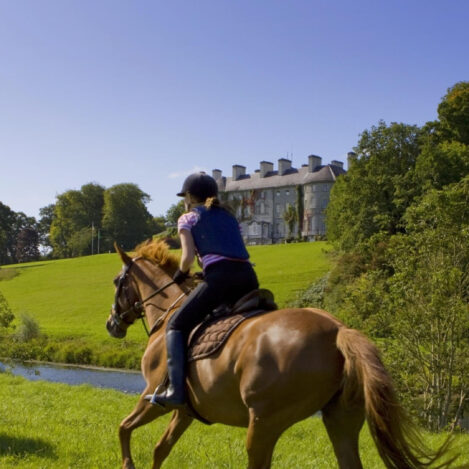 A couple horseback riding near a grand countryside hotel under a clear blue sky.