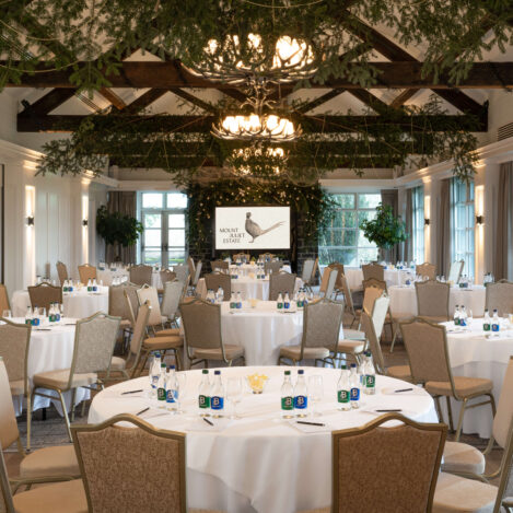 Elegant event hall with round tables set for guests, adorned with greenery and natural light.