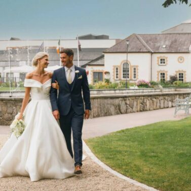 A couple walks joyfully on a garden path near a historic hotel, with a fountain in the background.